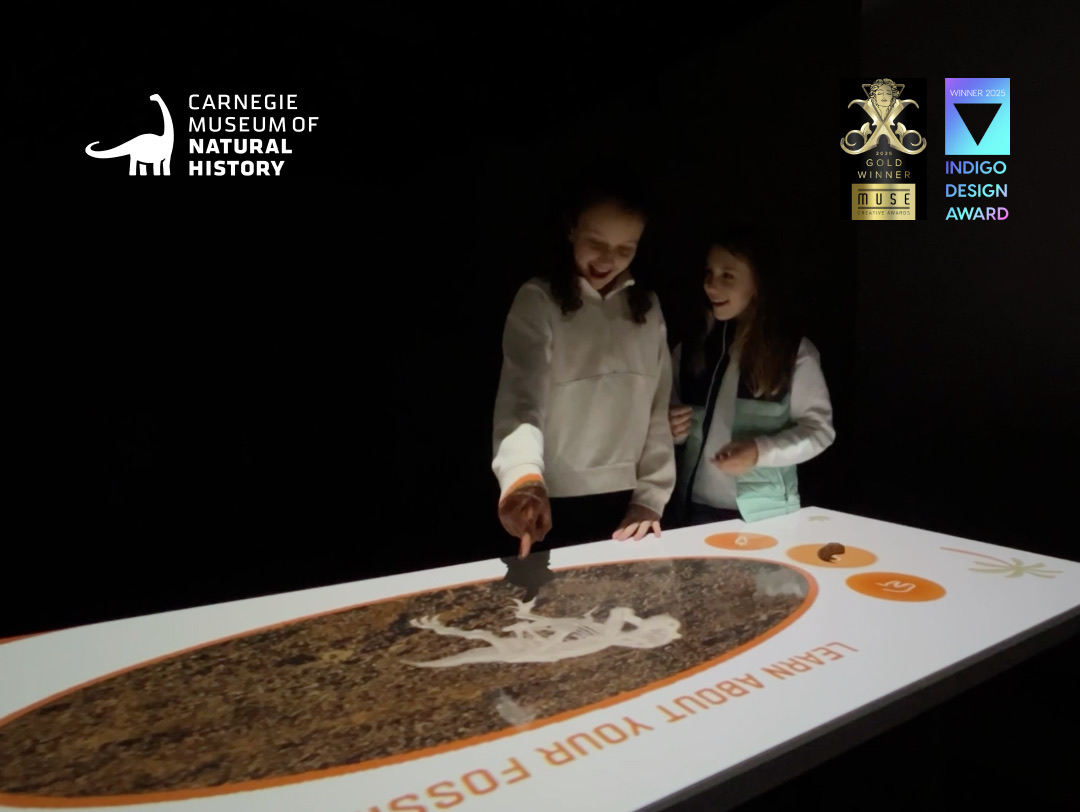 Two children digging for a fossil on an interactive sand table at the Carnegie Museum of Natural History, with award badges
